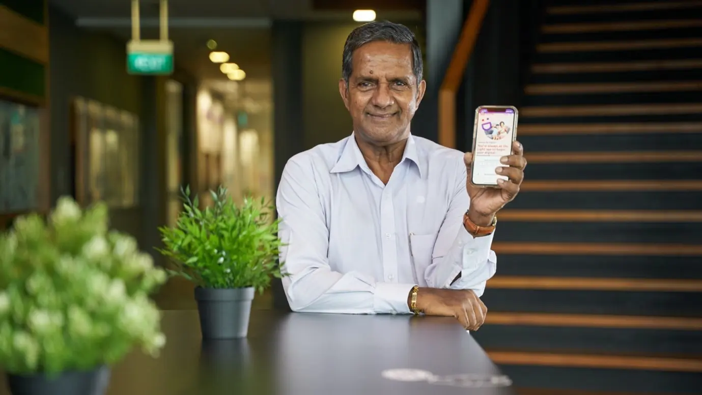 An elderly man smiles at the camera while holding up his phone, promoting digital literacy through the Digital for Life movement