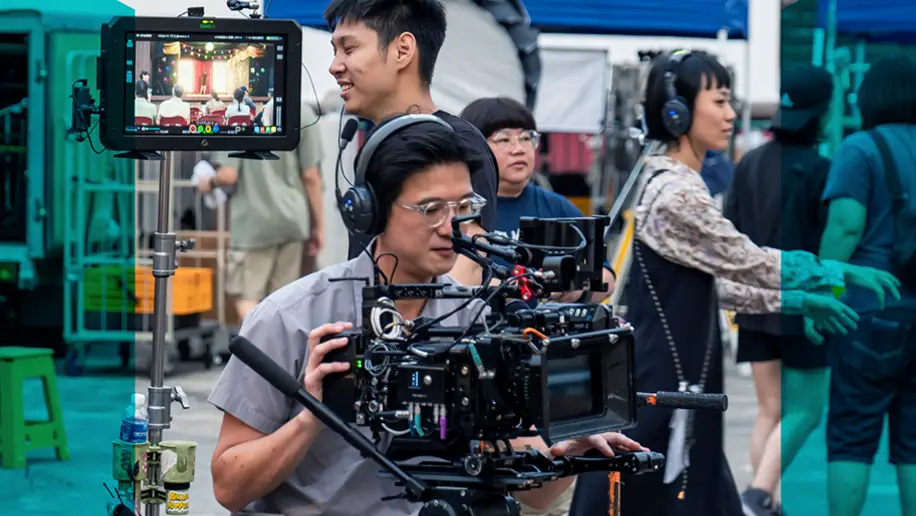 In the foreground, a videographer is checking the video camera on the set of Wonderland, a Made with SG film. Production crew and equipment are in the background, including a camera monitor, and a producer wearing a headset walking past. 