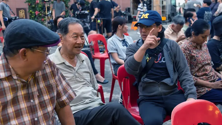 Actors Johnny Ng and Zhang Wei, and Director Chia Yee Wei on the set of Wonderland, a Made with SG film. They are seated in an outdoor setting, with the rest of the actors and crew in the background getting ready or waiting to shoot a scene.