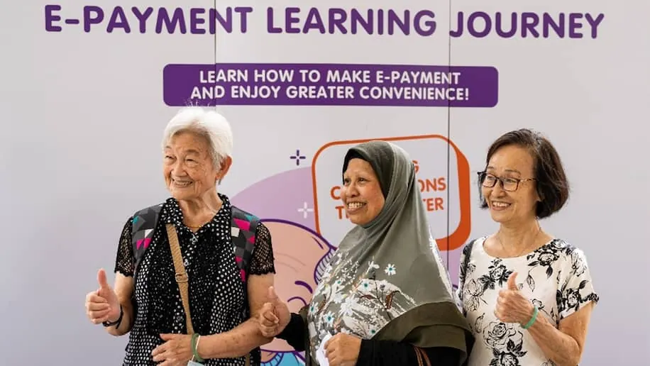 A group picture of three senior citizens posing by a backdrop for an e-payments learning workshop and doing a thumbs up sign.