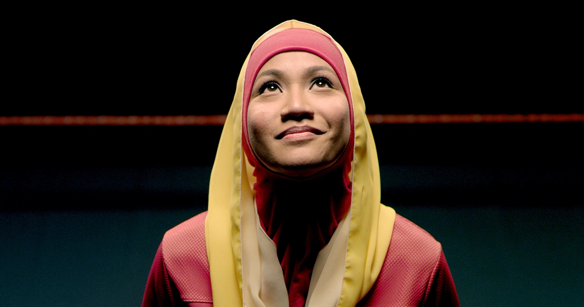 A film still from "Banting" showing a woman in a hijab looking upward with a hopeful expression against a dark background.