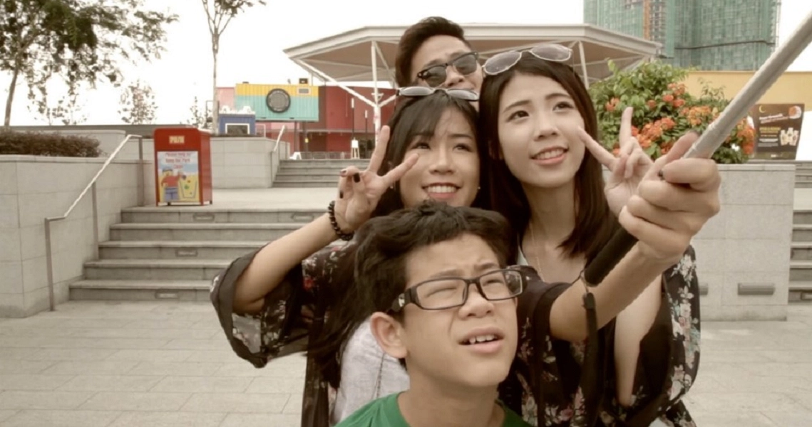 Film still from "Ariel and Olivia" showing three people smiling and gesturing outdoors near concrete steps and urban buildings.