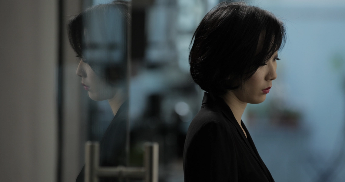 A woman in a dark suit stands in profile, her reflection visible in a window, in a film still from Lang Tong.