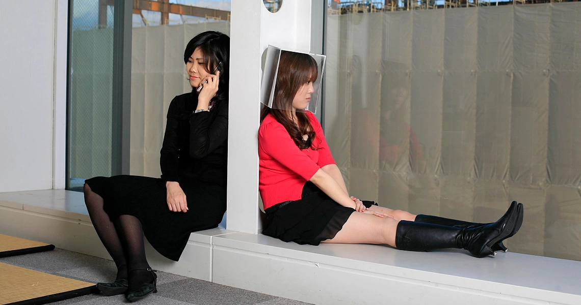Film still from Hashi showing two women sitting separately on a white ledge, one in black on phone, other in red sweater.