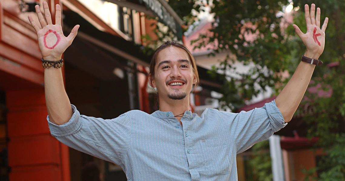 A smiling young man in a light blue shirt raises his hands in celebration outside colorful buildings in a scene from the film "The Leap Years".