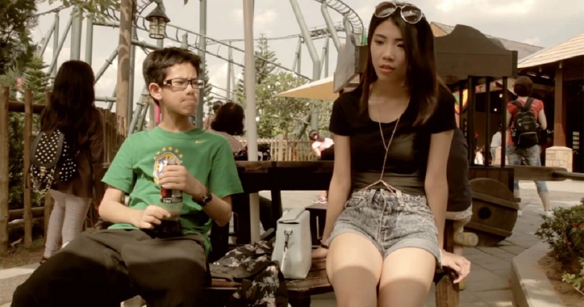 A scene from the film "Ariel and Olivia" showing two young people sitting at an outdoor cafe table, one wearing a green shirt.