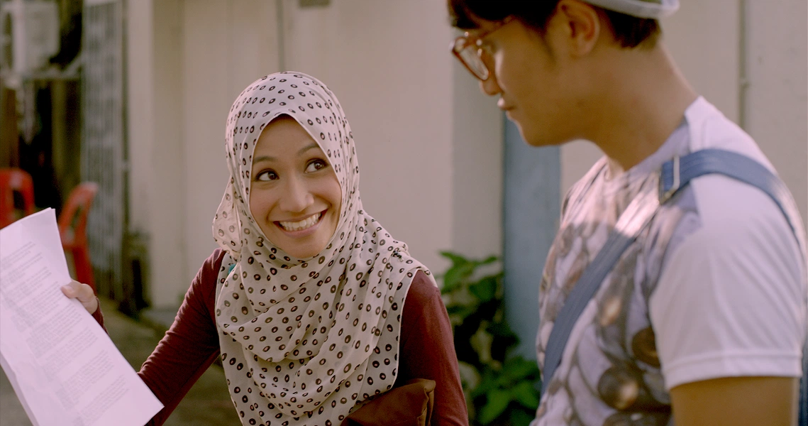 A scene from the film Banting showing a smiling woman in hijab holding papers and interacting with a man wearing glasses.